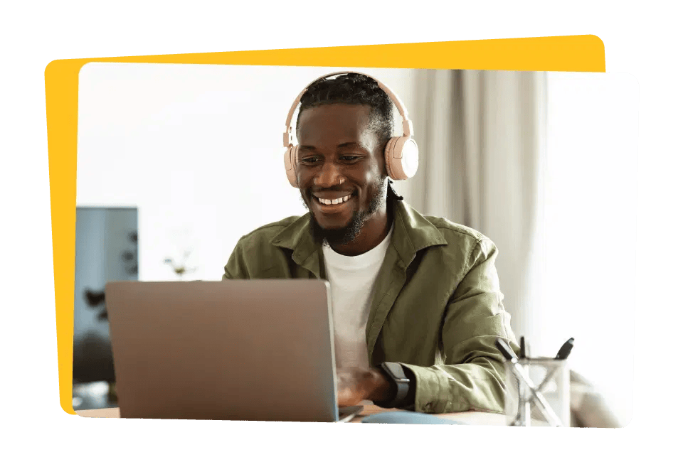 A smiling man wearing headphones uses a laptop at a desk in a brightly lit room.