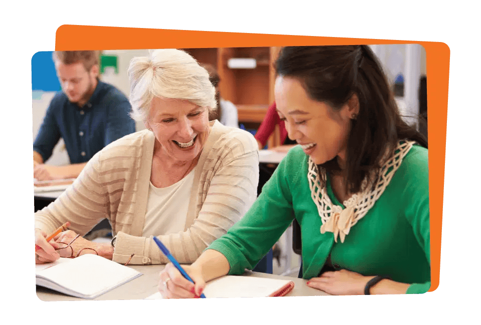 Two women, one older and one younger, smiling and looking at a notebook together in a classroom setting.