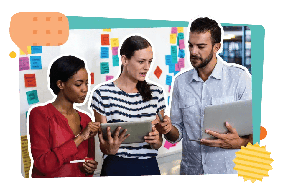 Three colleagues discuss work using a tablet and laptop in front of a colorful sticky note-covered wall.