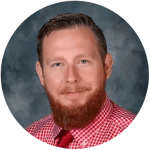 A person with a reddish-brown beard and short hair wearing a red and white checkered shirt and red tie against a gray background.