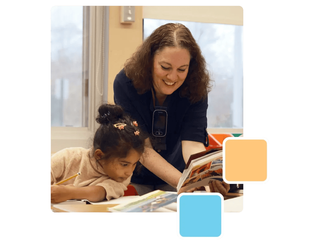 An adult helps a child read a book at a table. The child is writing, and the adult is smiling. The setting resembles a classroom, with windows in the background.
