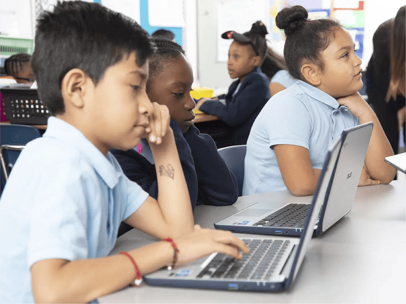 Three children sit at a classroom table, each using a laptop, with other students visible in the background.