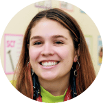 A young woman with long brown hair smiles at the camera, wearing hoop earrings and a lanyard, with colorful posters on the wall behind her.