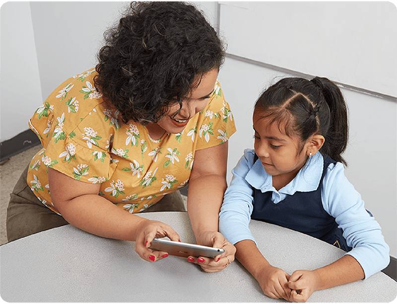 A woman shows a young girl something on a smartphone while they sit together at a table in a classroom, exploring reading comprehension strategies.